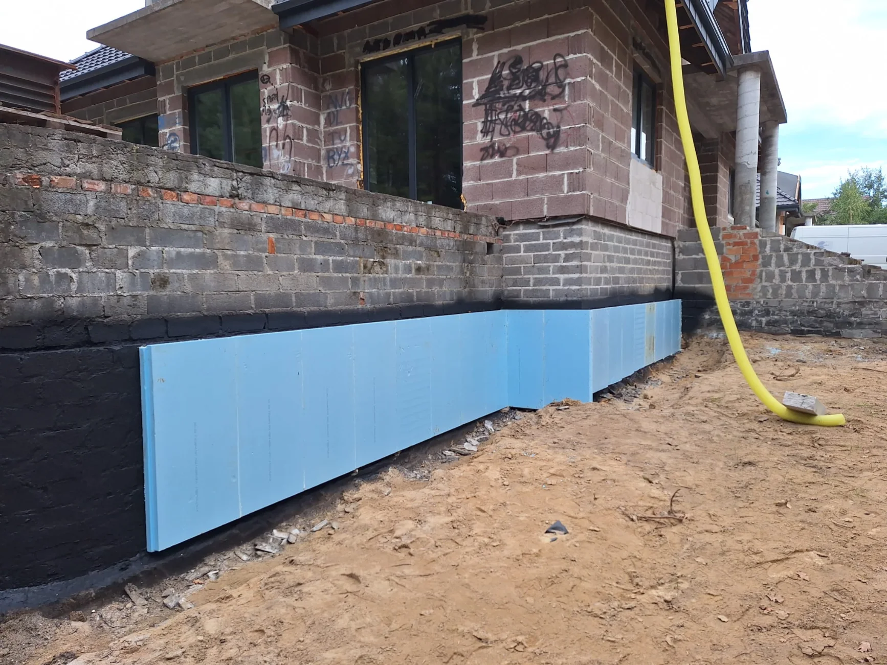 Izolacje fundamentów – montaż niebieskich płyt styropianowych przy murze domu w budowie